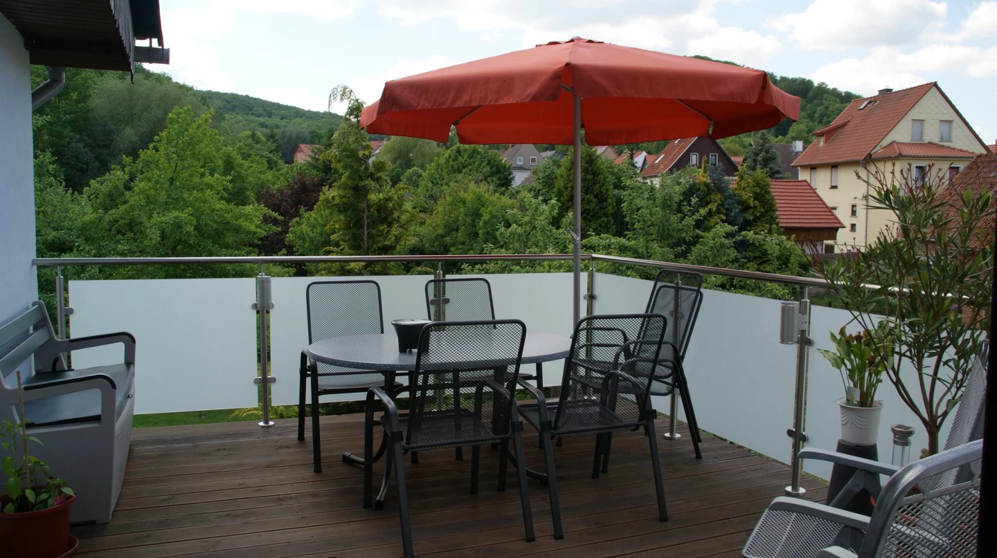 Holzbalkon mit Edelstahlpfosten und satiniertem Glas, Gartenmöbel unter rotem Sonnenschirm, dahinter Häuser und Wald