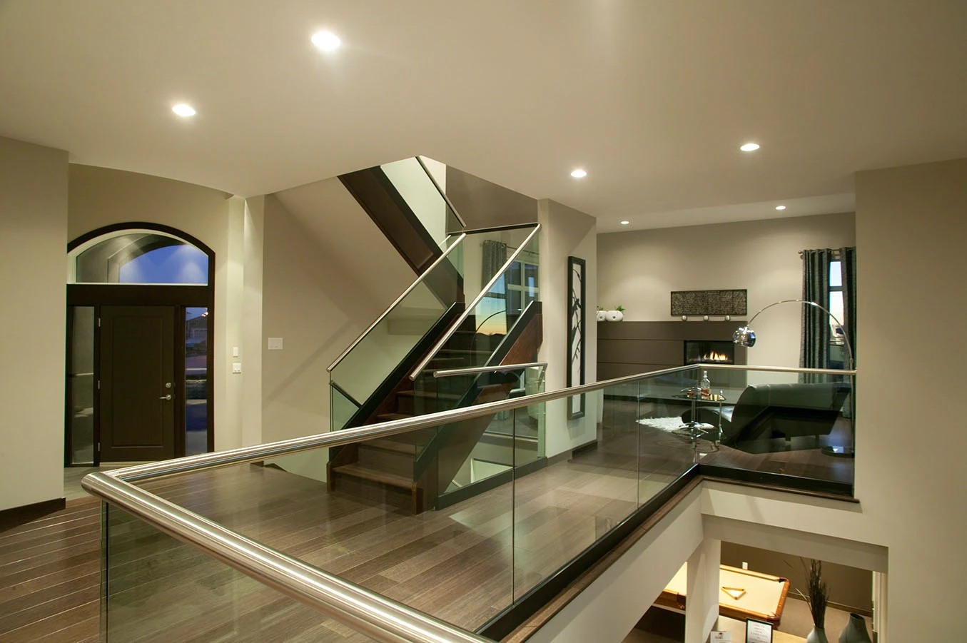 Innen-Galerie mit Glasbrüstung und rundem Edelstahl-Handlauf, dahinter eine Treppe mit Holzstufen und Glasgeländer, Blick in ein Wohnzimmer mit Kamin